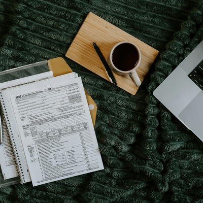 Tax documents and a binder on a desk beside a laptop and coffee